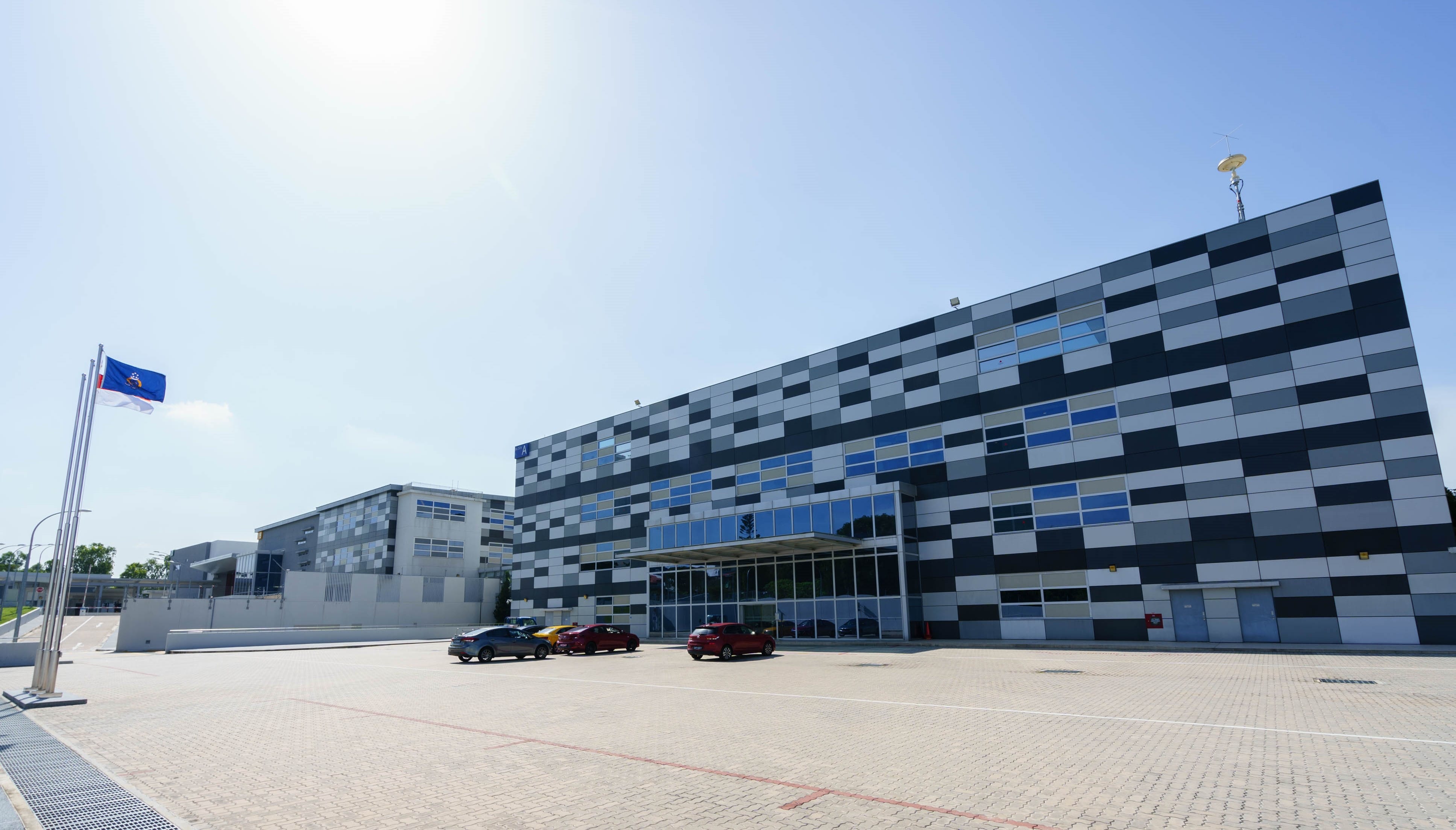 A large checkered building with parked cars and flagpoles stands in a sunny parking lot under a clear sky.
